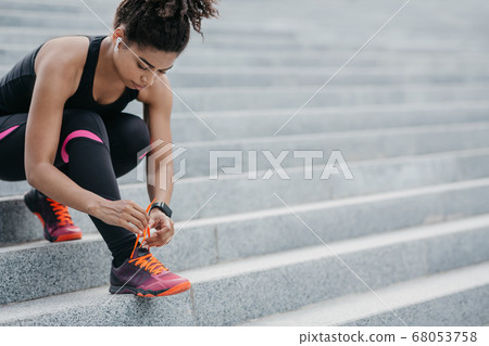 Runner getting ready for training. African american girl with wireless headphones and fitness tracker tying laces on sneakers 68053758