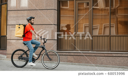 Delivery, work and sport. Young guy with beard in helmet with backpack rides a bicycle down the street 68053996