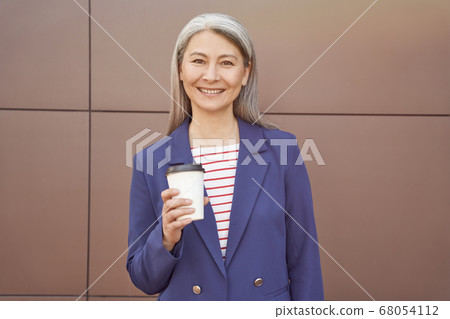 Coffee pause. Beautiful cheerful middle aged business woman wearing eyeglasses and classic suit holding cup of coffee and smiling at camera while standing against the wall outdoors 68054112