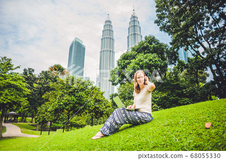 A young woman in casual dress using laptop in a tropical park on the background of skyscrapers. Mobile Office concept 68055330