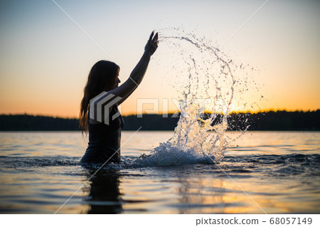 Beautiful Russian girl in a long black swimsuit swims outside the city on the lake in the rays of sunset or dawn. Splashing water on the background of the sun. Happy girl. Beautiful Russian girl in a long black swimsuit swims outside the city on the lake in the rays of sunset or dawn. Splashing water on the background of the sun. Happy girl. 68057149