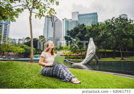 A young woman in casual dress using laptop in a tropical park on the background of skyscrapers. Mobile Office concept 68061755