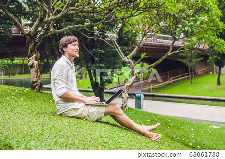 Man businessman or student in casual dress using laptop in a tropical park on the background of skyscrapers. Dressing in a white shirt, beige shorts. Mobile Office concept Man businessman or student in casual dress using laptop in a tropical park on the background of skyscrapers. Dressing in a white shirt, beige shorts. Mobile Office concept 68061788