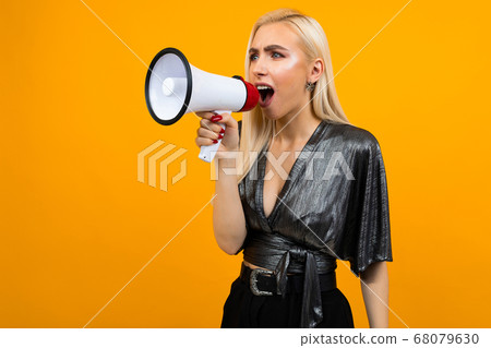 portrait of a caucasian blond girl in a graphite blouse talking news in a megaphone on a yellow studio background portrait of a caucasian blond girl in a graphite blouse talking news in a megaphone on a yellow studio background 68079630