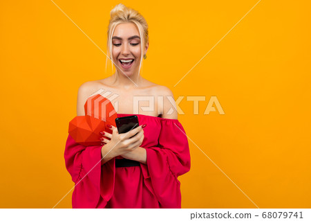 sad caucasian blond girl in a red dress communicates on a dating site with a phone and a red heart made of paper in hands on an orange studio background sad caucasian blond girl in a red dress communicates on a dating site with a phone and a red heart made of paper in hands on an orange studio background 68079741