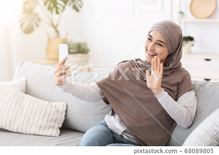 Video call. Muslim girl in hijab waving to smartphone camera at home 68089805
