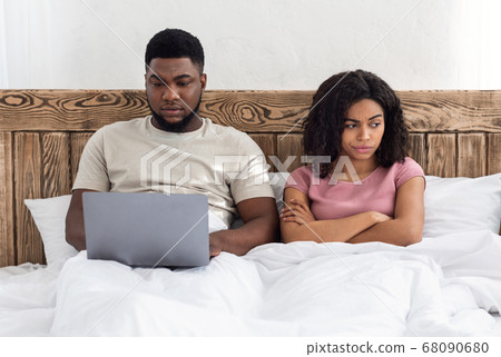 Addicted black man using laptop in bed, woman looking angry 68090680