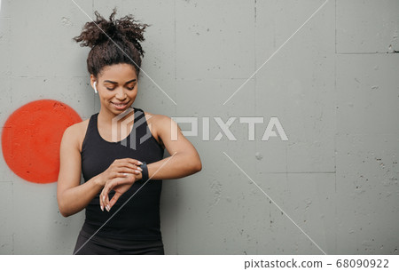 Jogging result. Happy african american girl with wireless headphones checks pulse on fitness bracelet Jogging result. Happy african american girl with wireless headphones checks pulse on fitness bracelet 68090922