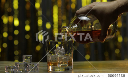 Pouring of golden whiskey, cognac or brandy from bottle into glass with ice cubes. Shiny background Pouring of golden whiskey, cognac or brandy from bottle into glass with ice cubes. Shiny background 68096386