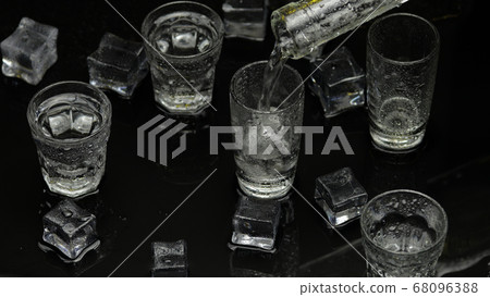 Barman pour frozen vodka from bottle into shot glass. Ice cubes against dark wet black background Barman pour frozen vodka from bottle into shot glass. Ice cubes against dark wet black background 68096388