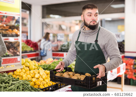 Positive man employee showing box with kiwi in grocery shop 68123662