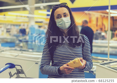 woman in protective medical mask in a supermarket chooses cheese. pandemic and covid-19 concept. Hand of the buyer with a piece of cheese in the store. toned woman in protective medical mask in a supermarket chooses cheese. pandemic and covid-19 concept. Hand of the buyer with a piece of cheese in the store. toned 68125255