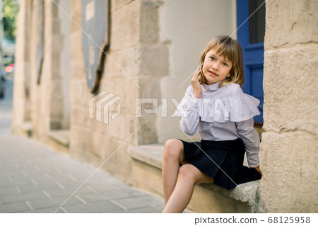 Outdoor fashion street city portrait of little Caucasian girl. Happy little girl sitting near the old building wall in ancient European city with blue door on the background Outdoor fashion street city portrait of little Caucasian girl. Happy little girl sitting near the old building wall in ancient European city with blue door on the background 68125958