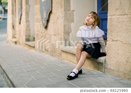 Outdoor fashion street city portrait of little Caucasian girl. Happy little girl sitting near the old building wall in ancient European city with blue door on the background 68125960