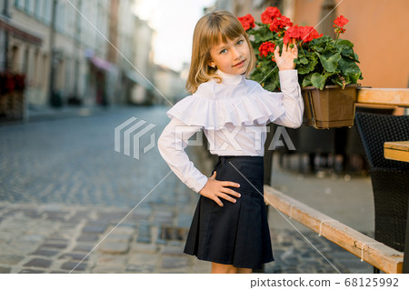 Closeup of little pretty blond Caucasian girl touching red flower pots decorations on summer day in old ancient European city 68125992