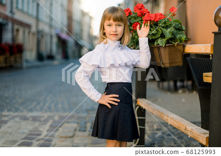 Closeup of little pretty blond Caucasian girl touching red flower pots decorations on summer day in old ancient European city 68125993