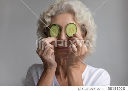 Elderly woman covers eyes with cucumbers slices enjoy skincare procedure 68131028