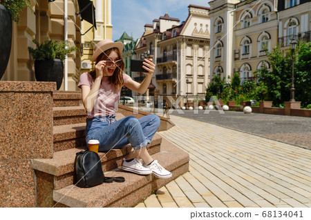 Beautiful Young Woman tourist with takeout coffee sitting on stairs using smartphone Beautiful Young Woman tourist with takeout coffee sitting on stairs using smartphone 68134041
