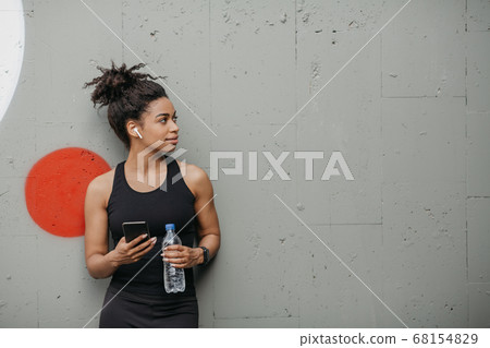 Muscular african american girl with modern gadgets for workout holds bottle and smartphone on gray wall background in city Muscular african american girl with modern gadgets for workout holds bottle and smartphone on gray wall background in city 68154829
