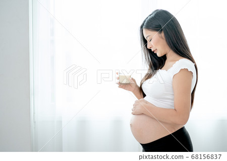 Beautiful Asian pregnant woman hold glass of milk and look to her belly in front white curtain Beautiful Asian pregnant woman hold glass of milk and look to her belly in front white curtain 68156837
