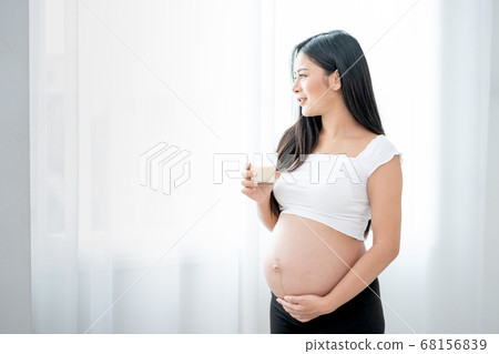 Beautiful Asian pregnant woman hold glass of milk and look outside through white curtain Beautiful Asian pregnant woman hold glass of milk and look outside through white curtain 68156839