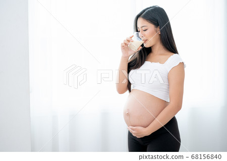 Beautiful Asian pregnant woman drink milk and stand in front of white curtain and she look happy Beautiful Asian pregnant woman drink milk and stand in front of white curtain and she look happy 68156840