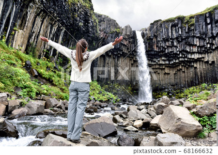 Iceland. Woman enjoying majestic Svartifoss waterfall. Female is visiting famous tourist attraction of Iceland. Spectacular natural landmark on vacation in Skaftafell. Icelandic nature landscape 68166632