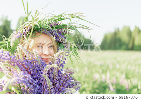 A blonde girl in a flower lupine wreath with a bouquet of lupines gently smiles and inhales the A blonde girl in a flower lupine wreath with a bouquet of lupines gently smiles and inhales the 68167370