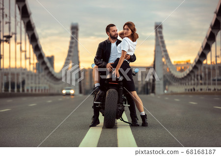 Young sexy woman hugging cute man in stylish black leather jacket, sitting on sports motorcycle on the bridge in the city on sunset and kissing 68168187