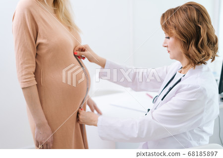 Serious female doctor wearing white coat measure belly waist size of pregnant young woman Serious female doctor wearing white coat measure belly waist size of pregnant young woman 68185897