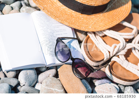 Beach hat on opened book with sunscreen and shoes on pebble beach Beach hat on opened book with sunscreen and shoes on pebble beach 68185930