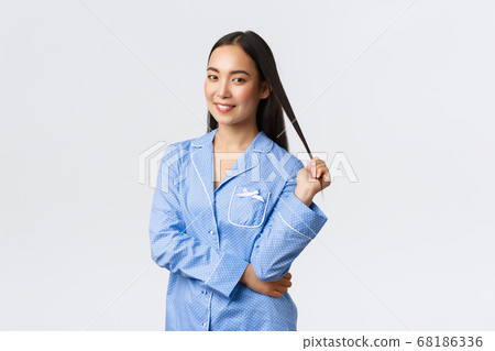 Tender gorgeous asian girl smiling dreamy at camera, standing in blue pajamas over white background. Woman prepare go to sleep in jammies, apply skincare products before bed 68186336