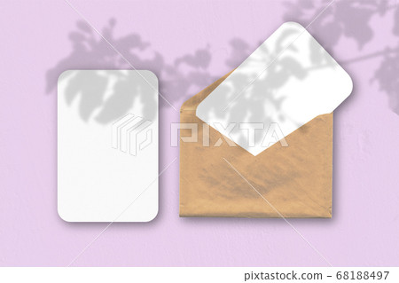An envelope with two sheets of textured white paper on a pink table background. Mockup with an overlay of plant shadows. Natural light casts shadows from a tropical plant. 68188497