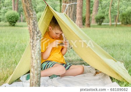 Boy playing video game outdoor in park, backyard, garden. Kid with phone in tent. 6 years old child having fun on nature. E-learning concept. Children and gadgets. Entertainment. Summer holidays Boy playing video game outdoor in park, backyard, garden. Kid with phone in tent. 6 years old child having fun on nature. E-learning concept. Children and gadgets. Entertainment. Summer holidays 68189082