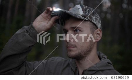 Portrait of handsome young man hunter or tourist. Man puts on his head cap with diode flashlight on visor in evening. 68190639