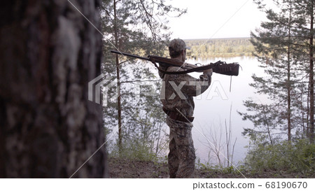 View from back of hunter in camouflage puts hunting rifle on his shoulder and looks at river from forest. Outdoor activity concept. Summer hiking in wilderness area. 68190670