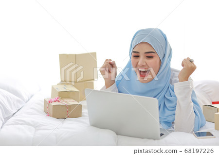 asian muslim woman in sleepwear with attractive, lies on bed with computer and online package box asian muslim woman in sleepwear with attractive, lies on bed with computer and online package box 68197226