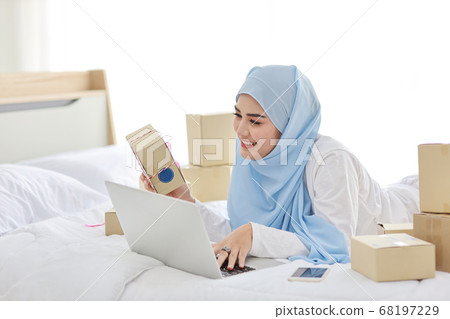 asian muslim woman in sleepwear with attractive, lies on bed with computer and online package box asian muslim woman in sleepwear with attractive, lies on bed with computer and online package box 68197229