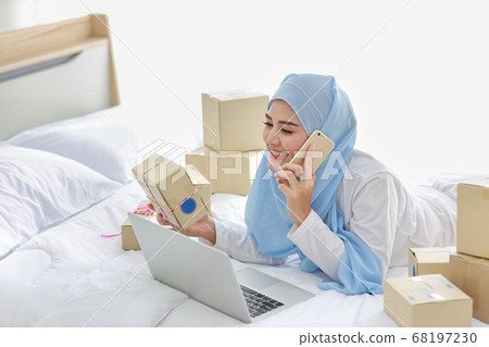 asian muslim woman in sleepwear with attractive, lies on bed with computer and online package box asian muslim woman in sleepwear with attractive, lies on bed with computer and online package box 68197230