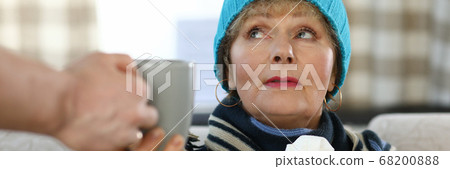 Elderly woman in scarf and hat sits on sofa home Elderly woman in scarf and hat sits on sofa home 68200888