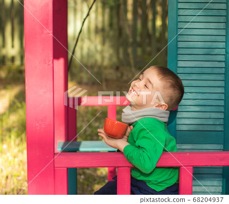 A laughing boy with a red Cup sitting on a wooden porch against the background of a green forest 68204937