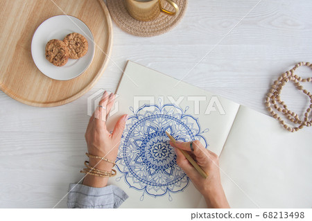 Female hands close up draw Decorative round floral mandala. Hobby and home relaxation. A mug of coffee and cookies on a wooden tray. White background. Female hands close up draw Decorative round floral mandala. Hobby and home relaxation. A mug of coffee and cookies on a wooden tray. White background. 68213498