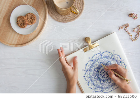 Female hands close up draw Decorative round floral mandala. Hobby and home relaxation. A mug of coffee and cookies on a wooden tray. White background. Female hands close up draw Decorative round floral mandala. Hobby and home relaxation. A mug of coffee and cookies on a wooden tray. White background. 68213500