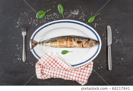 Whole smoked mackerel fish on white oval plate, gray black stone like table under, rock salt and green leaves salad near. View from above Whole smoked mackerel fish on white oval plate, gray black stone like table under, rock salt and green leaves salad near. View from above 68217841