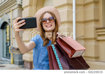 Young Woman with shopping bags making selfie walking in a city at summer day Young Woman with shopping bags making selfie walking in a city at summer day 68220019