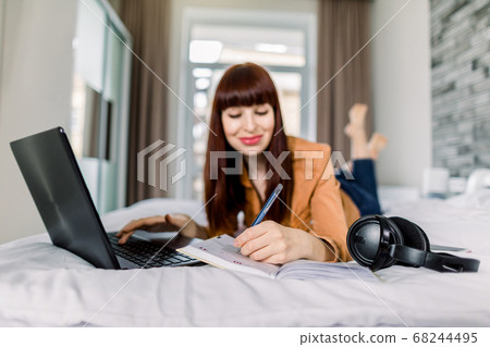 Happy smiling dark haired woman lefthander using laptop and making notes in her diary notepad, while lying on bed in the morning and watching video course. Focus on hand with pen 68244495