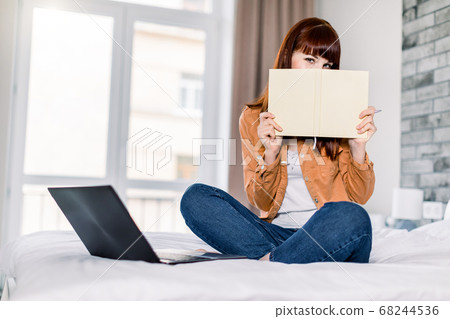 Happy funny beautiful woman in jeans and yellow casual shirt, hiding her face with notepad, while working on a laptop sitting on the bed at home. Freelance, work from home concept 68244536