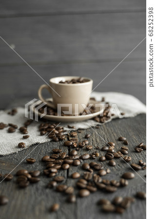 Coffee cup filled of fresh arabica or robusta coffee beans with scattered coffee beans on a linen textile and wood table. Coffee cup filled of fresh arabica or robusta coffee beans with scattered coffee beans on a linen textile and wood table. 68252398