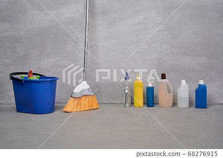 House cleaning and disinfection. Plastic bucket with cleaning products, broom, bottles with different detergents standing on the floor against grey wall 68276915