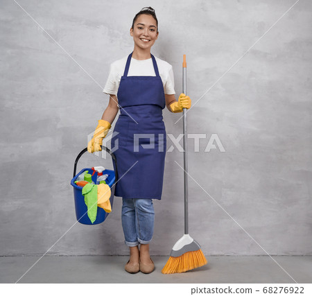 Happy cleaning lady in uniform and yellow rubber gloves holding broom and plastic bucket with rags and different cleaning products while standing against grey wall Happy cleaning lady in uniform and yellow rubber gloves holding broom and plastic bucket with rags and different cleaning products while standing against grey wall 68276922